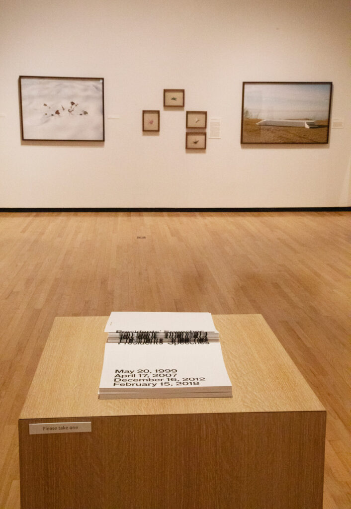 Image: Andres Gonzales. President’s Speeches, American Origami (his work from his series American Origami is in the background). Black and white pamphlets sit in a stack on a wooden pedestal in a gallery. Several other photographs hang on display on a wall in the background. Photo by Sarah Hoskins.