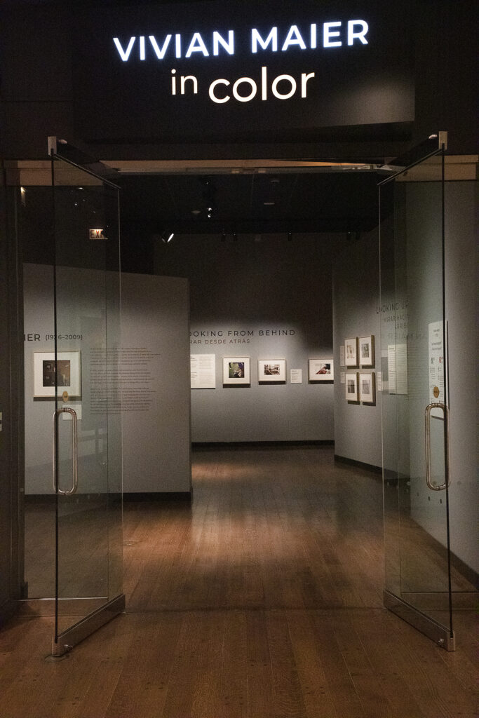 Image: A view of the exhibition Vivian Maier: In Color at the Chicago History Museum. The viewer's perspective is of outside the exhibition look in through two opened glass doors. Various photographs and exhibition text are on the grey exhibition walls. Photo by Sarah Hoskins.