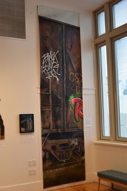Image: Stef Ratliff. "Train to Nowhere," 2021, acrylic on canvas, 168” x 43.5”. The painting on display hangs vertically almost from floor to ceiling. Mostly dark in color, the composition resembles the side of a train car with graffiti. Photo by Kim Kobersmith.