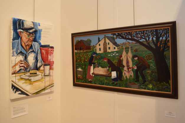 Image: Two paintings are displayed on a gallery wall. On left: Lacy Hale. "Papaw Breakfast," 2021, acrylic on canvas, 24″ x 36″. The painting shows a man wearing glasses and a hat eating breakfast. On right: Nan Phelps. "Butchering Hogs," 1985, oil on Masonite. The painting shows a country scene of several people butchering a hog. A house and a barn are in the background. Photo by Kim Kobersmith. 