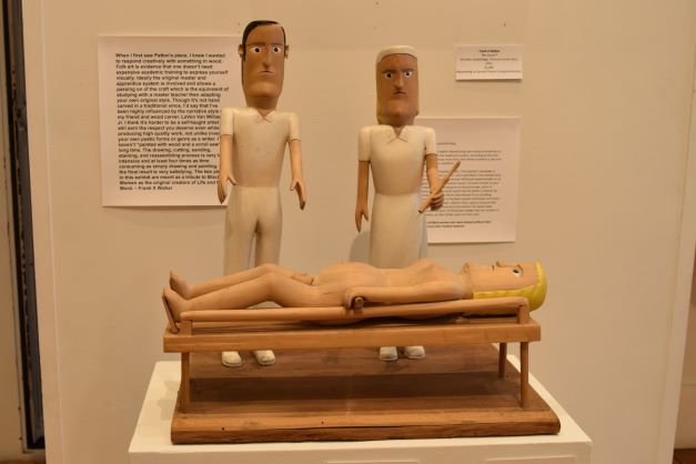 Image: Ernest Patton. "Hospital Birthing," 1989, paint and wood. A sculpture comprised of two wooden figures in white that stand in front of a third wooden figure. The third figure is nude and laying down. Photo by Kim Kobersmith.