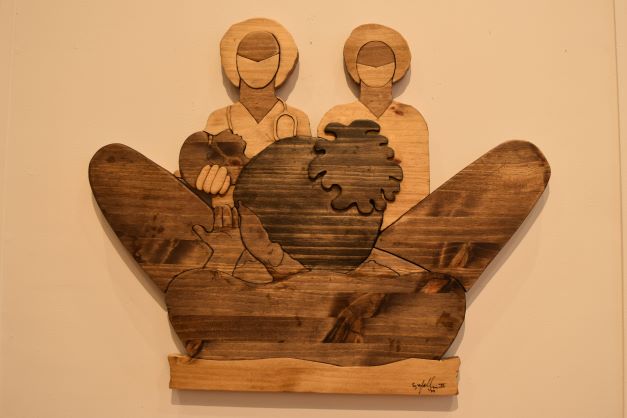 Image: Frank X Walker. "No Doula?," 2021, wooden assemblage, pine and wood stains. The piece on display is made out of carved wood and shows a woman giving birth in the foreground and two doctors standing in the background. Photo by Kim Kobersmith.