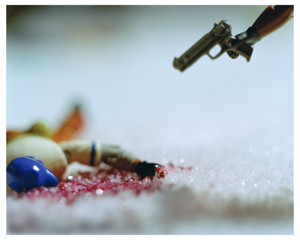 Image: Hank Willis Thomas and Kambui Olujimi. "Still from Winter in America," 2005. A video still showing a toy person laying down in snow wounded from a gunshot. A toy hand comes out from the right side of the frame and holds a toy gun pointed at the toy person. Courtesy of the artists and Jack Shainman Gallery.