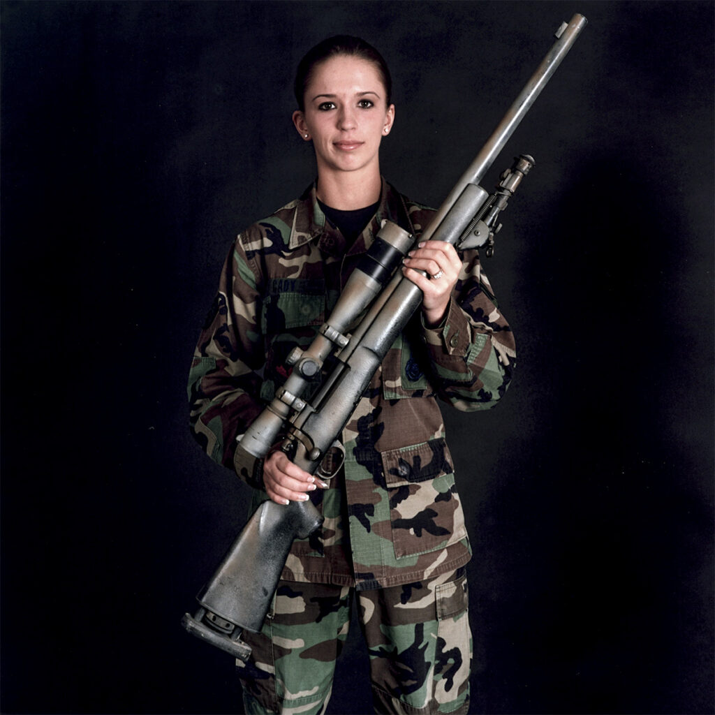 Image: Nancy Floyd. "A1C Ashley-Ann Cady with M24 (Remington 700 bolt action rifle), Moody Air Force Base, Georgia," 2006. A photograph of a woman in a camouflage outfit stands in front of a black background while holding a gun. She looks directly at the camera. Courtesy of the artist.