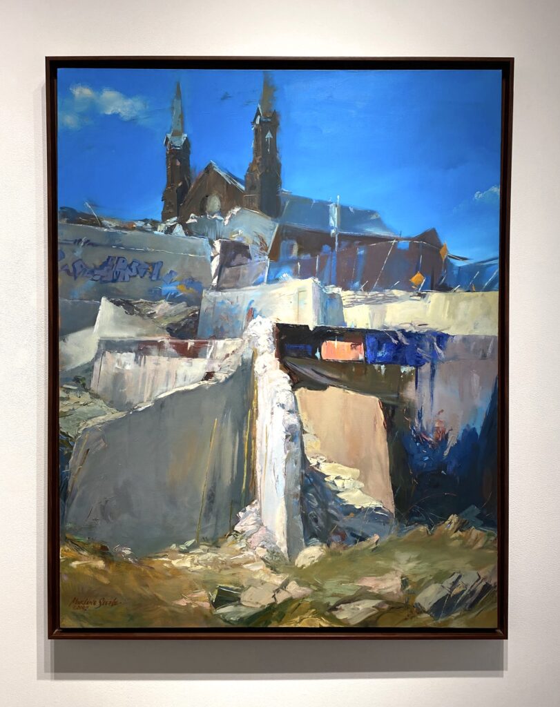 Image: Marlene Steele, "Reflections in Ruins," 2007, oil on canvas. A painting of a church-like building with concrete blocks in front of it hangs on a white wall. Photo of artwork by the author.