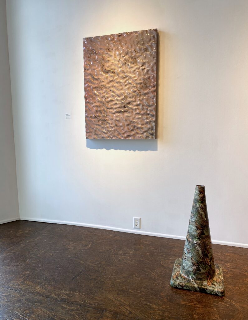 Image: Two pieces by Collin Richard on display at Manifest Gallery. The two dimensional, textural piece hanging on the wall is "Growth of the Soil," 2021, resin, MDF, Miracle-Gro. The sculptural, cone-like piece sitting on the gallery floor is "Wayfinder," 2021, hydrographic film print on traffic cone. Photo by the author.