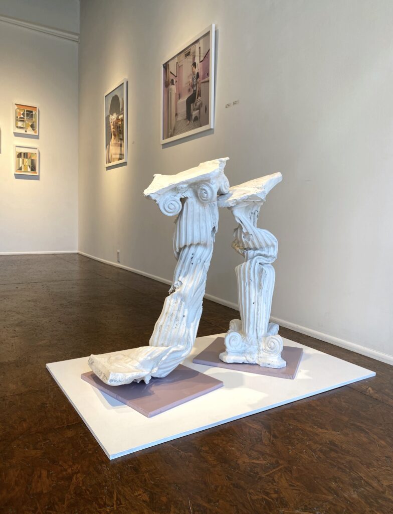 Image: A view of the exhibition Post-Urban at Manifest Gallery. The sculptural piece in the foreground by Sarah Deppe looks similar to two ionic columns that have been bent and broken. The piece is titled Persephone & Rhea, 2021, plaster, foam and wood. The works on display in the background are by Rebecca Harrell and Jesse Egner. Photo by the author.