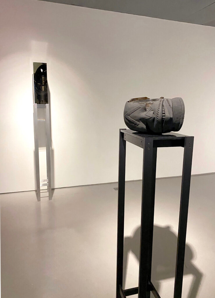 Image: Matthew Angelo Harrison: (L) Seer: Peering through Aurora, 2020, headlight, tinted polyurethane resin, anodized aluminum, acrylic. (R) Dark Silhouette: Synthetic Lipiko no. 5, 2018, wooden sculpture from West Africa, polyurethane resin, anodized aluminum, acrylic. Photo by Ely Maris. Two sculptures sit across from each other in a gallery. The piece in the foreground sits on a dark pedestal and somewhat resembles a human head. The piece in the background is dark in color, rectangle in shape, and sits on a light-colored pedestal.