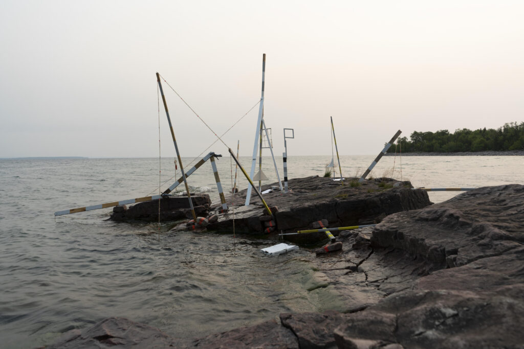 Image: Claudia O’Steen and Aly Ogasian, Keweenaw Observing Station (19 Jul, 8:33 PM), 2021, giclée print. An photo of a site-responsive, portable sculpture created by the artists, located on a rocky shore of Lake Superior with trees in the background. Image courtesy of Manifest Gallery.