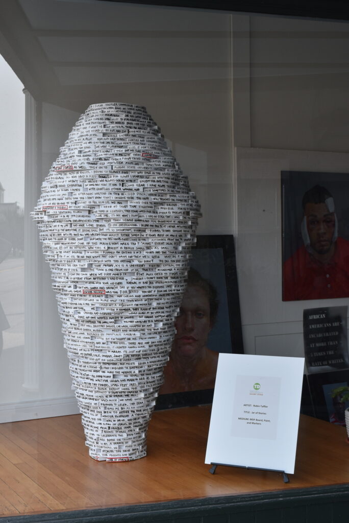 Image: Robin Taffler, Jar of Stories, 38” x 60”, MDF Board, paint, markers. A mostly black and white sculpture sits on a wooden surface. The sculpture resembles a vessel and is covered completely in text. Photo by Kim Kobersmith.