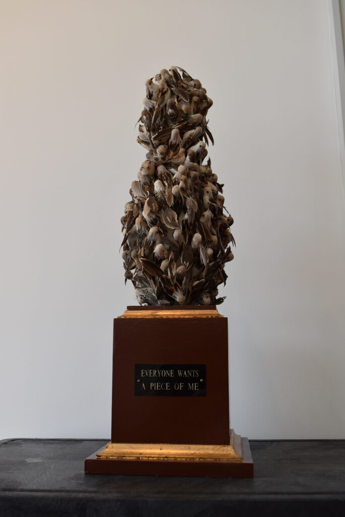 Image: Robin Taffler, Everyone Wants a Piece of Me, 9” x 9” x 26”, mixed media. A mixed-media sculpture comprised of many small birds, which form a bowling pin-like shape. The piece has a pedestal base not unlike a trophy. The name of the piece is on a plaque on the base. Photo by Kim Kobersmith. 
