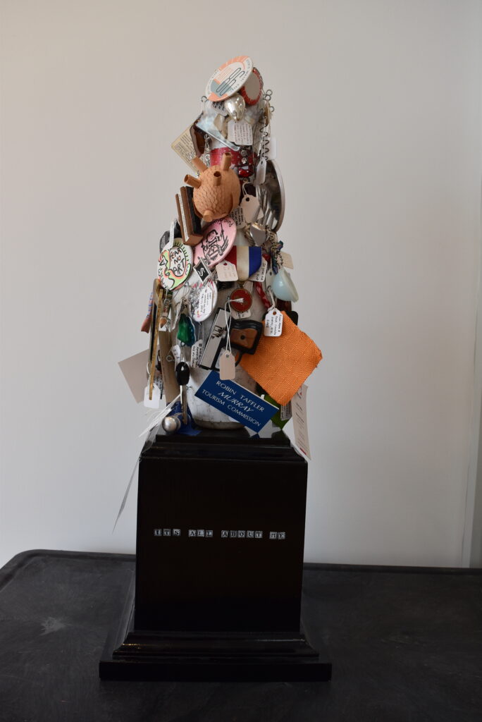 Image: Robin Taffler, It’s All About Me, 9” x 9” x 26”, mixed media. A bowling pin shapes sculpture on a black pedestal. The piece is covered in personal tchotchkes like ticket stubs and name tags.