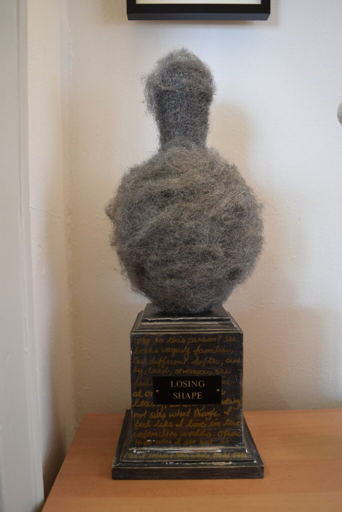 Image: Robin Taffler, Losing Shape, 9” x 9” x 26”, mixed media. A sculpture comprised of grey fuzz on a a pedestal base that is not unlike a trophy. The name of the piece is on a plaque on the base. Photo by Kim Kobersmith.