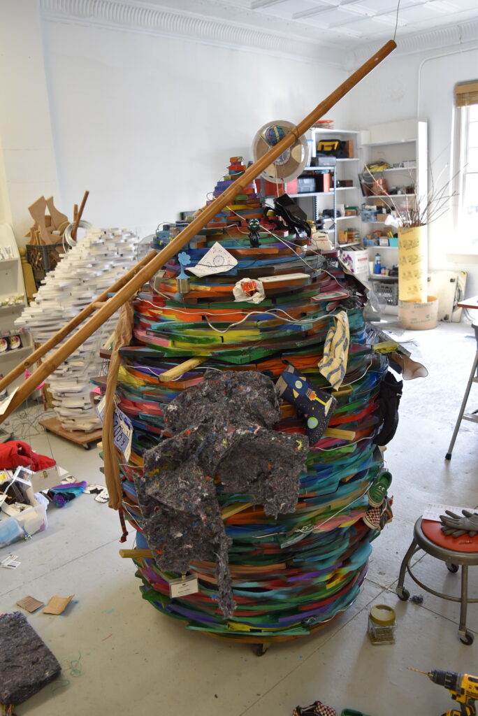 Image: Robin Taffler, Big Chaos, work in progress. A large, colorful, mixed-media sculpture in the shape of an elongated sphere. The sculpture sits in the artist's studio space. Photo by Kim Kobersmith.