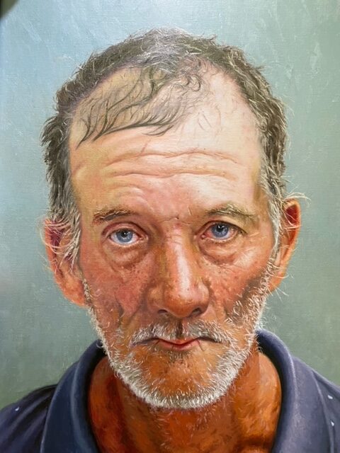 Image: Dick Dougherty, Arrested: S-102Q19/621, 2022, oil on linen covered panels, 24” x 36”. A painting of a portrait of a man looking straight into the camera for a mug shot. He has white hair, a beard, blue eyes, and wears a blue colored shirt. Photo provided by the artist.