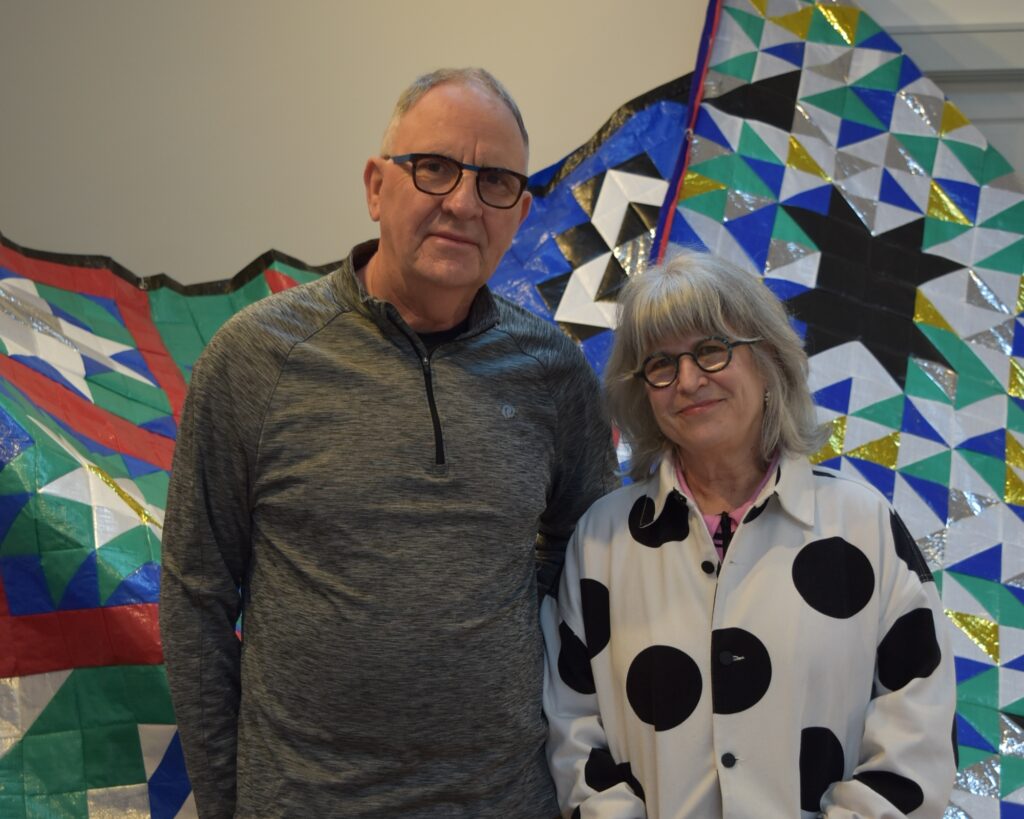 Image: Dick Dougherty and Robin Taffler stand in front of a quilt-like background. Dougherty, on the let, wears black glasses and a grey shirt. Taffler, on the right, wears round glasses and a black and white polkadot shirt. Photo by Kim Kobersmith.