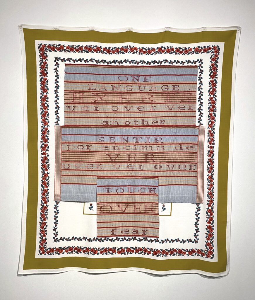 Image: Hellen Ascoli, Touch Over Fear, 2020, handwoven cotton and wool, hand-stitched on found fabric, 56" x 49". Photo by Ely Maris. A rectangle textile piece hangs on a white wall. It reads: "ONE LANGUAGE EXERTS ver over ver another SENTIR por encima de VER over ver over TOUCH OVER fear." The border is made up of an olive green solid line and small roses.
