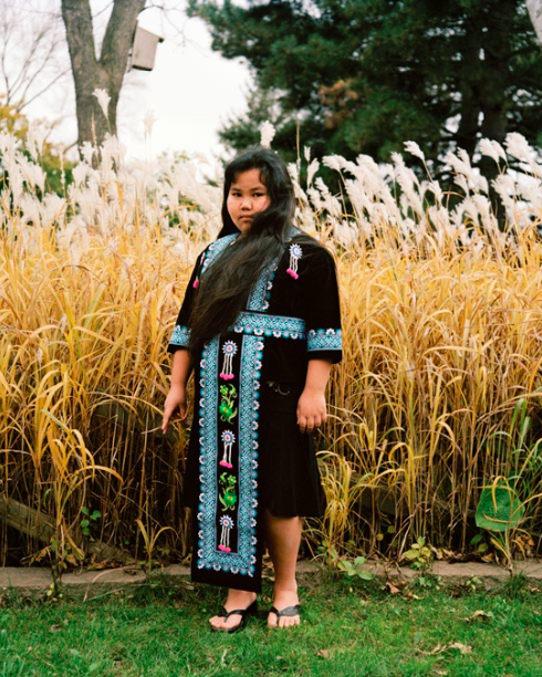 Image: Pao Houa Her, Mai Youa in Hmong Clothes, 2006-09, digital archival inkjet print, 40" x 32". Photo of the work is by Ely Maris. A photograph of a young girl in traditional Hmong clothes standing in front of wheat field. She looks directly at the viewer.