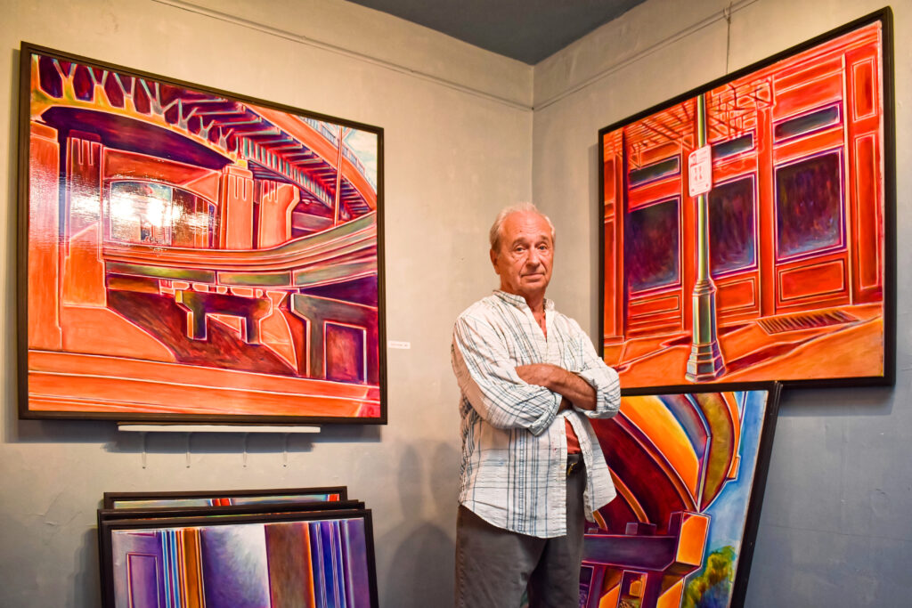 Image: A photo of Clay Wainscott standing in front of two of his paintings hanging on the wall and several of his paintings leaning up against the wall. He is looking straight at the viewer with his arms crossed. Photo by Kevin Nance.