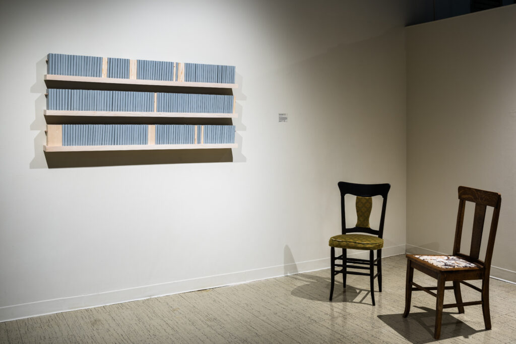 Image: Becky Alley, Epitaphs for Edwards, 2016, 220 artists books each with a hand-stitched epitaph inside. An installation in a gallery of two wooden chairs on the right, and three shelves holding light blue and beige books on the left. Image courtesy of the artist.