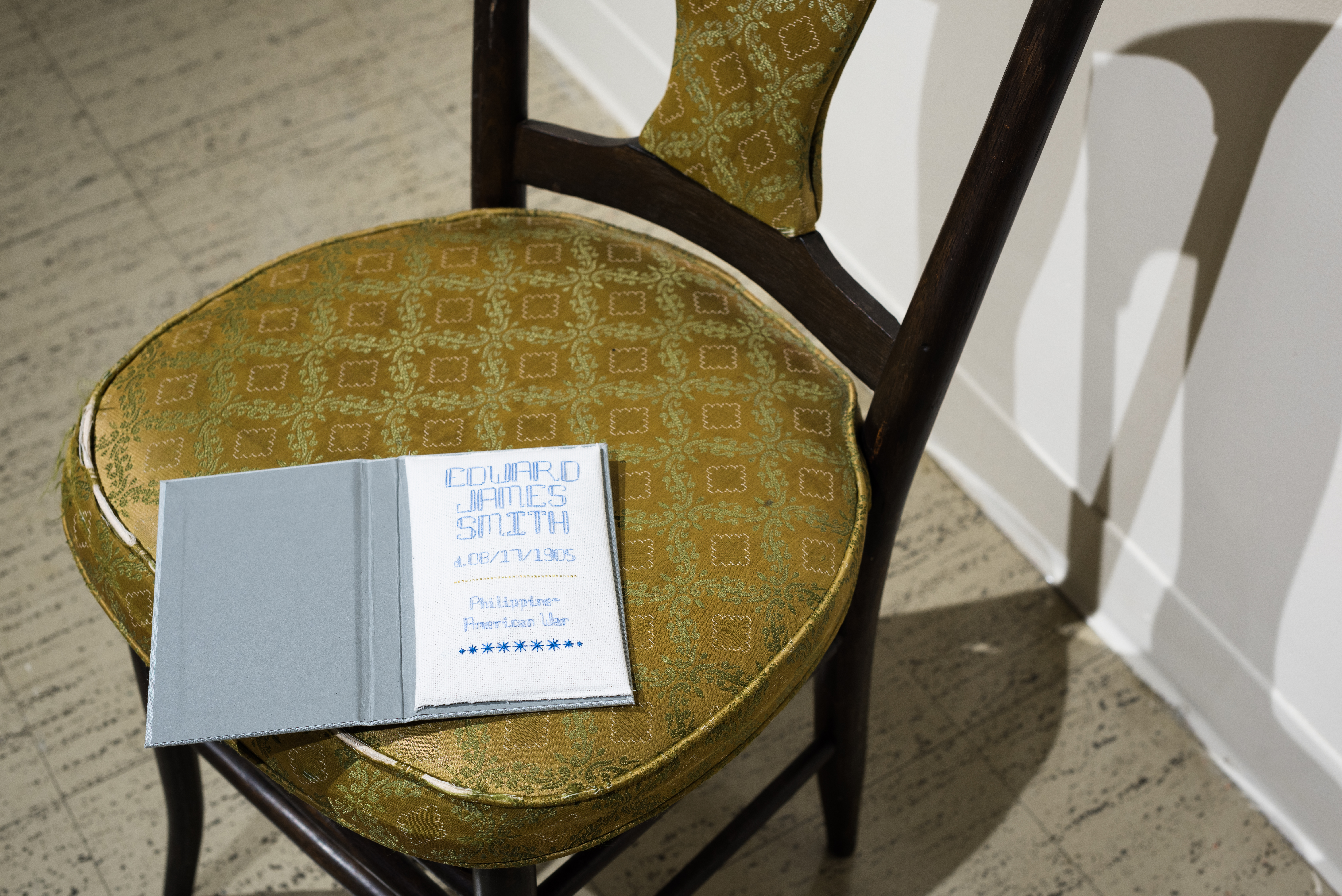 Image: Becky Alley, Epitaphs for Edwards, 2016, 220 artists books each with a hand-stitched epitaph inside. A detail shot of the installation showing one wooden chair with a gold-colored cushion. Sitting on the chair is a light blue book open so that you can see the first page, which reads: "COWARD JAMES SMITH". Image courtesy of the artist.