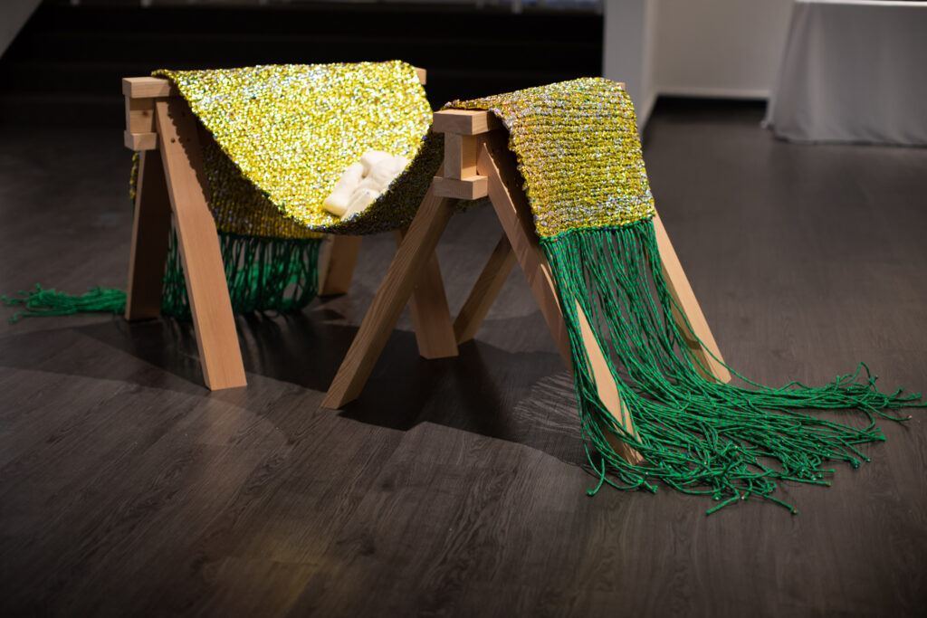 Image: Becky Alley, Heirlooms​, 2021, felted objects, woven emergency blankets, paracord, cedar saw horses. An installation of a yellow textile hanging over cedar saw horses. The piece has green tassels hanging down on either side. Image courtesy of the artist.
