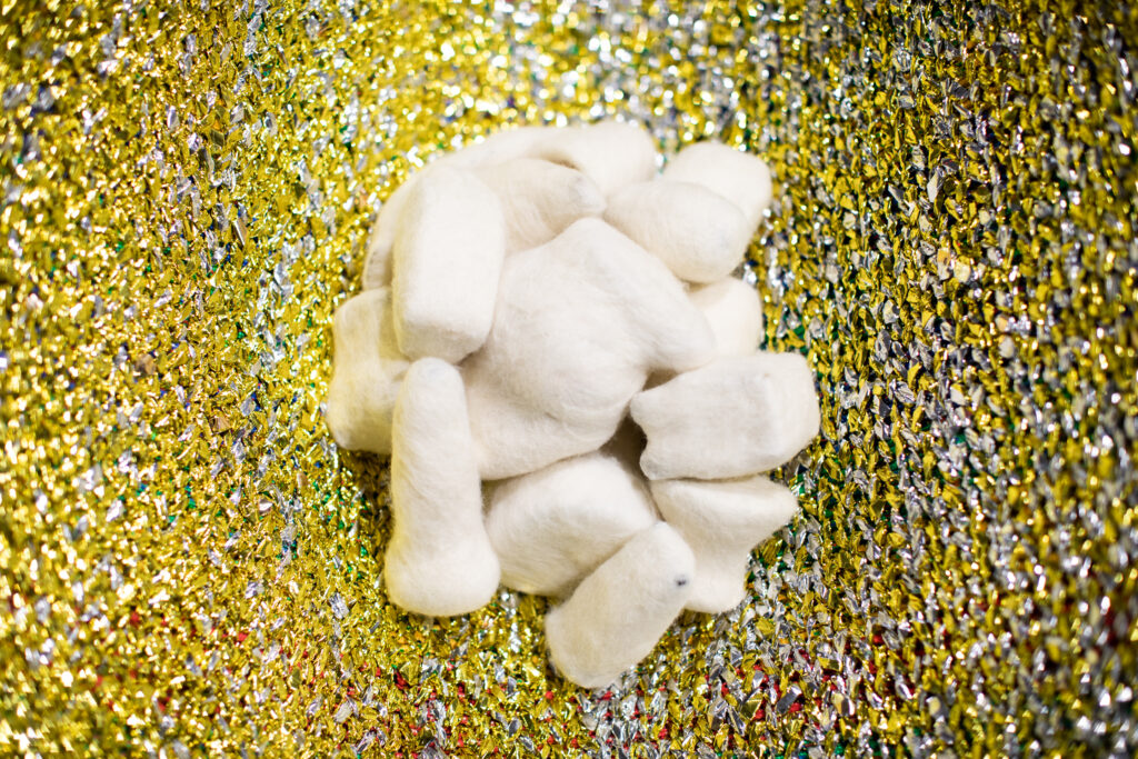 Image: Becky Alley, Heirlooms​, 2021, felted objects, woven emergency blankets, paracord, cedar saw horses. A detail shot of Heirlooms, showing white felt pieces clumped together on a mostly yellow textile piece. Image courtesy of the artist.