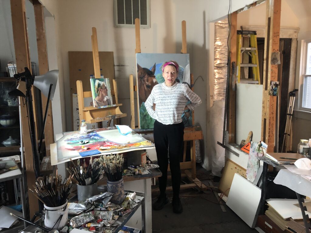 Image: Gaela Erwin in her studio, 2022. The artist stands in her studio looking directly at the viewer. Surrounding her are various painting materials such as paint brushes, paint palettes, paper, and studio lights. Behind the artist are several paintings propped up on easels. Photograph by Peter Morrin. 