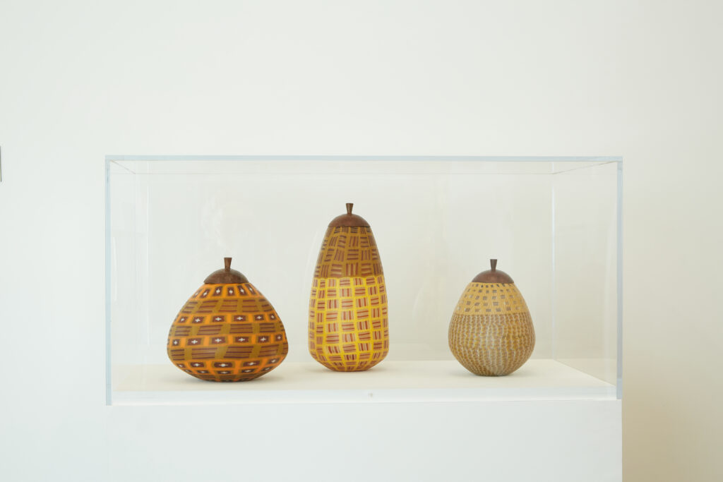 Image: Corey Pemberton, Ukhambas (fall), 2021, brown glass, steel, patina. Dimensions variable. Three orange, yellow, and brown vessels are on display in a glass case. Courtesy of the artist and KMAC Museum. Photo by Ted Wathen.