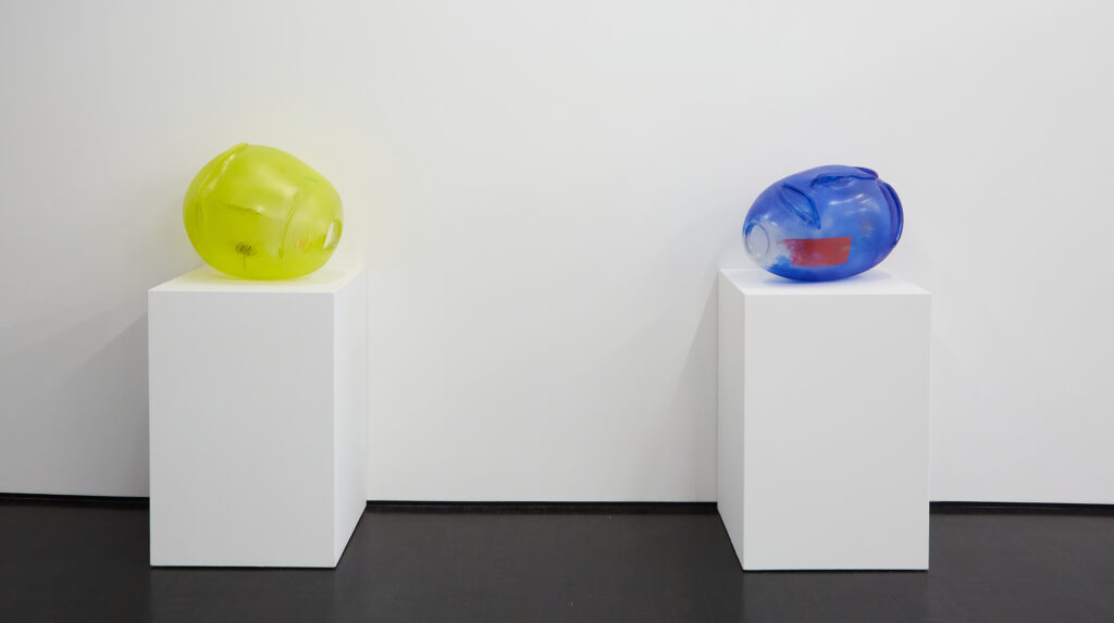 Image: Therman Statom, Sol, and Aqua Fuego, 2017, blown glass and pigment, 22" x 19", 24" x 16". Two round glass sculptures are on display on white pedestals. The one on the left is yellow on the one on the right is primarily blue. Courtesy of the artist and KMAC Museum. Photo by Ted Wathen.