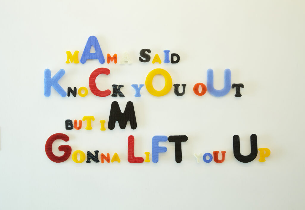 Image: SaraBeth Post, Remix #1, 2021, cast glass, 38" x 66" x 2". Multi-colored letters spell out "MAMA SAID KNOCK YOU OUT BUT IM GONNA LIFT YOU UP" on a white wall. Courtesy of the artist and KMAC Museum. Photo by Ted Wathen.