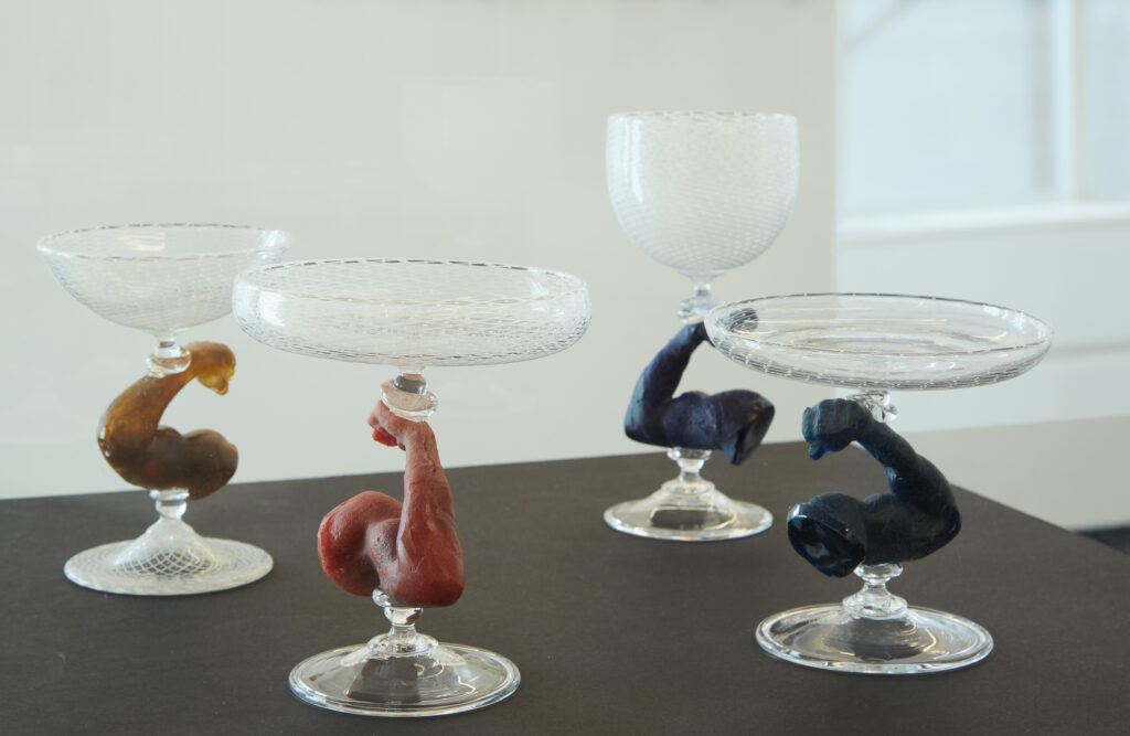 Image: Ché Rhodes, Flex, 2021, blown and cast glass, variable dimensions. Four glasses with stems that look like arms that are flexing sit on a dark table. Courtesy of the artist and KMAC Museum. Photo by Ted Wathen.