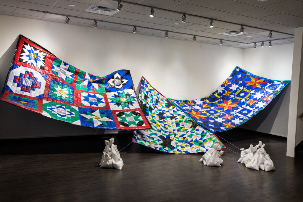 Image: Becky Alley, Quilts​, 2021, tarps, emergency blankets, bedsheets, paracord, sand. An installation in a gallery comprised of several hanging quilts. The patterns are mostly green, white, blue, yellow, and red and are made up of geometric shapes. The quilts are being held up by cords that connect to white sandbags that sit on the gallery floor. Image courtesy of the artist.