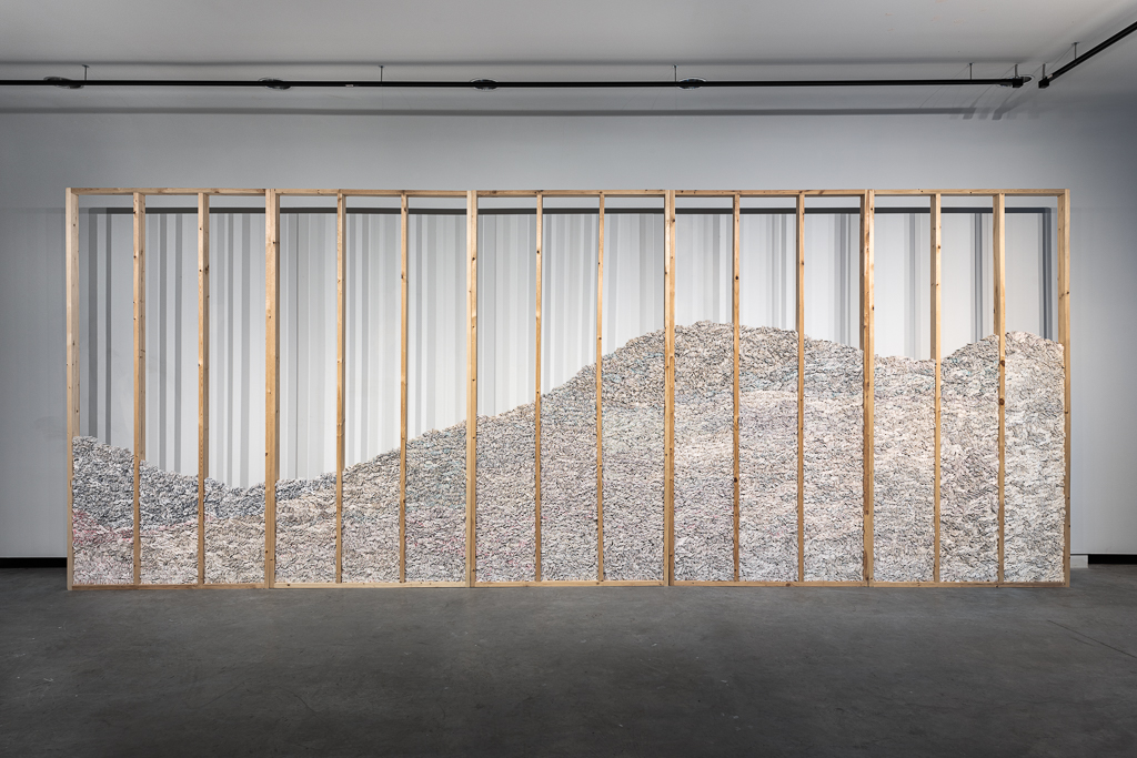 Image: Becky Alley, Rubble, 2018 (ongoing), 120,000 white flags: used bed sheets, plaster; pine. A war memorial for civilians killed in Syria since 2011. The photo shows the piece installed in a gallery. Several vertical planks of wood form a cage-like structure holding in a giant pile of white bed sheets. Image courtesy of the artist.