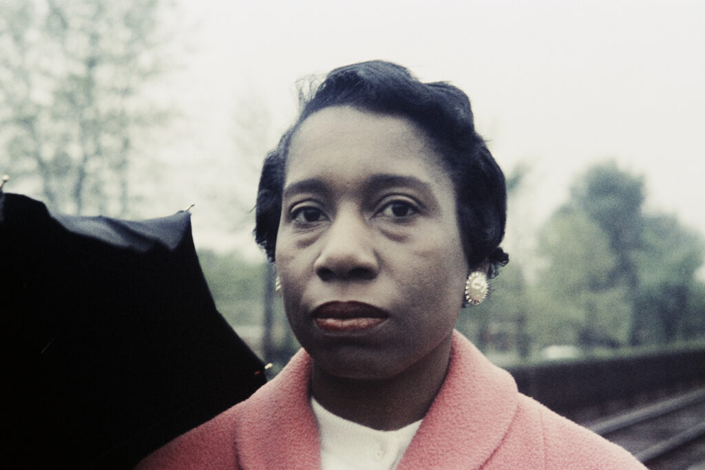 Image: Vivian Maier, Portrait of a woman in a pink coat. Likely Chicago suburbs, c.1956-74.Chicago History Museum, ICHi-180976 / © The Estate of Vivian Maier. A photograph of a Black woman with short hair wearing a pink coat. She is looking directly at the viewer.