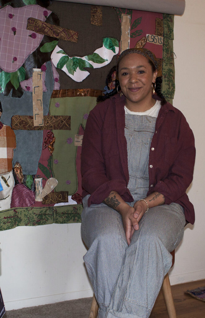 Image: Interdisciplinary artist Josie Love Roebuck in her home/studio in Villa Hills, Kentucky, March 2022. She is sitting with her hands in her lap and smiles at the viewer. One of her large textile pieces hangs on a wall behind her. Photo by CM Turner.
