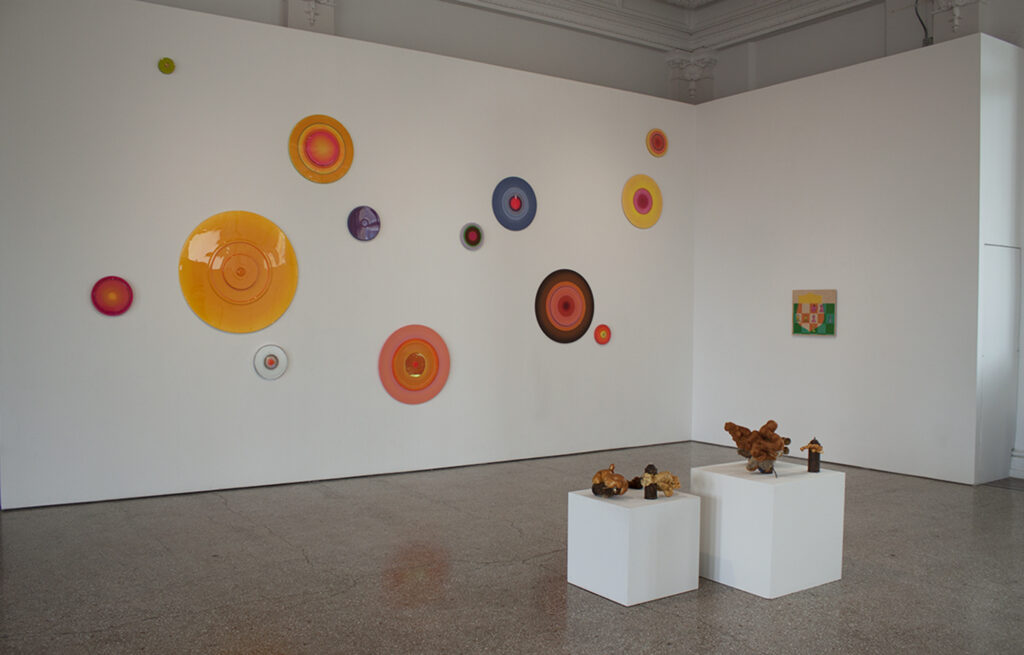 Image: An installation view of A Thought is a River, curated by John Knuth. Works on display are by Letitia Quesenberry, Matt Coors, and Albertus Gorman (Clockwise from Left). Several small sculptures are on display on pedestals in the foreground. On the wall in the background hangs one painting and a variety of colorful disk-shaped wall sculptures. Photo by CM Turner.