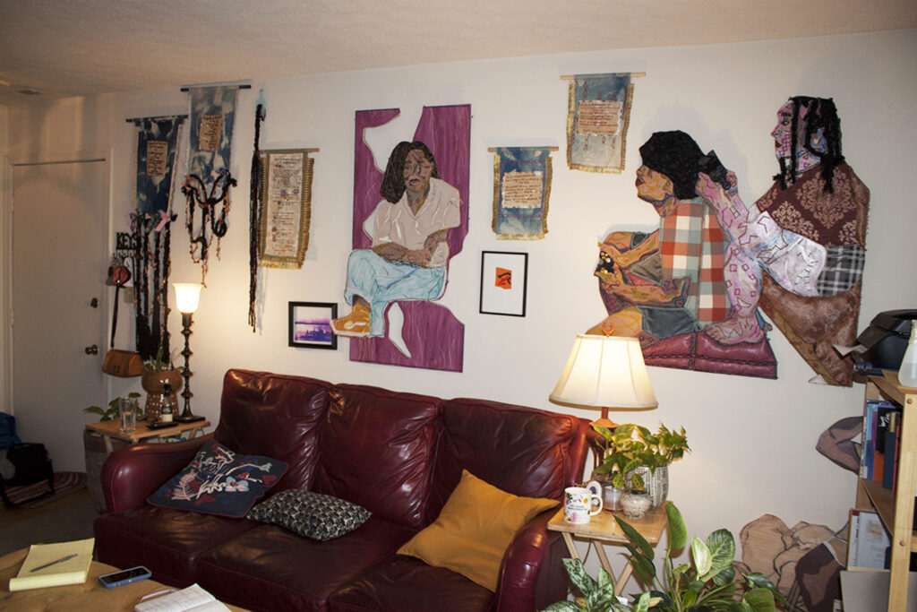 Image: An interior view of Josie Love Roebuck’s home/studio. Various textile works by the artist are on display on the wall, many of which depict people. Photo by CM Turner.
