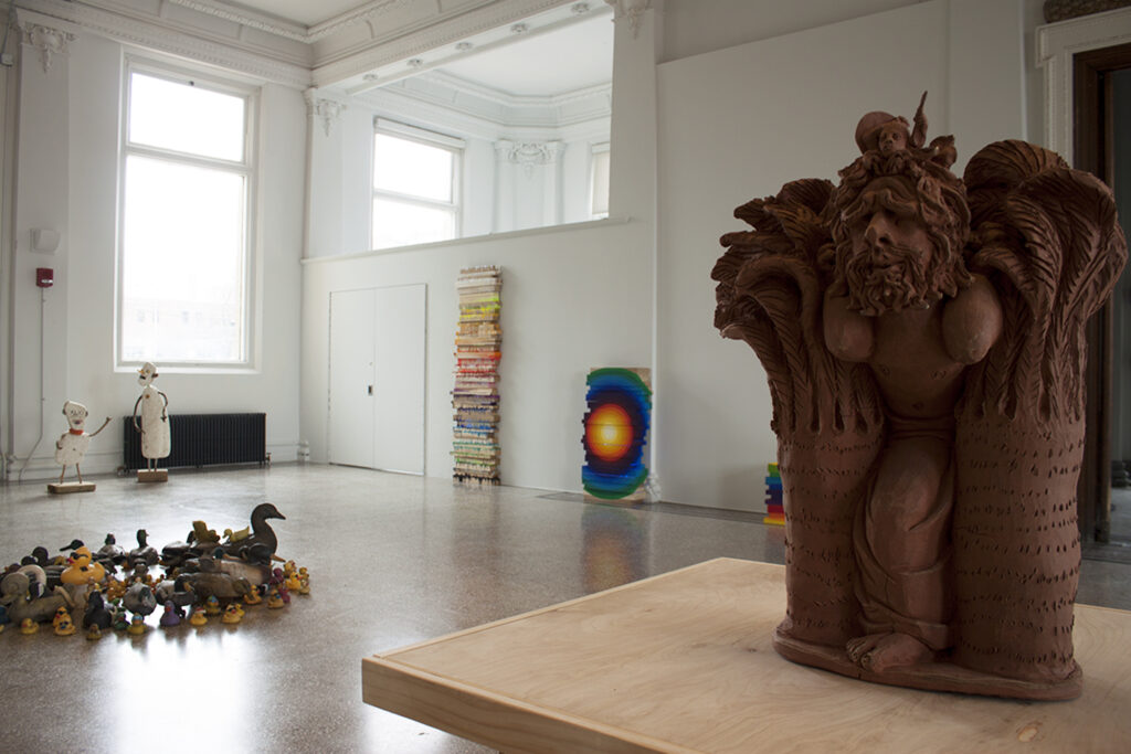 Image: An installation view of A Thought is a River, curated by John Knuth. Works on view in The Carnegie’s Rotunda Gallery are by Albertus Gorman, Adrienne Dixon, and Chris Hammerlein (Left to Right). In the foreground to the right is a dark brown sculpture on a wooden surface. In the background are several colorful sculptures leaning against the wall. To the left, wooden duck decoys and rubber duckies are arranged in a circle on the floor. Photo by CM Turner.