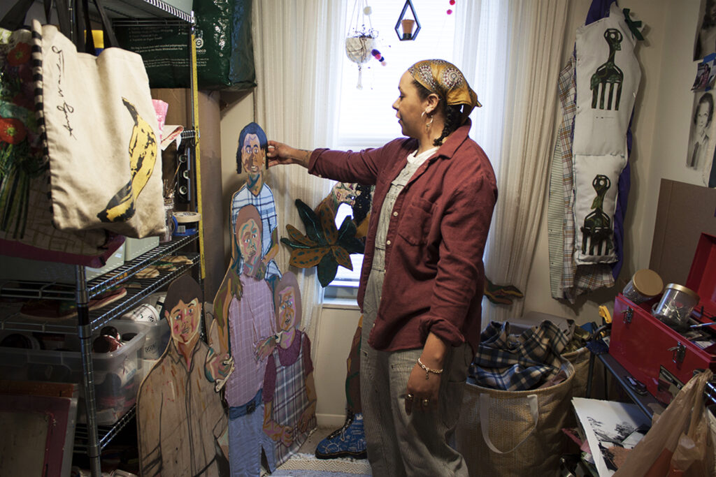 Image: Interdisciplinary artist Josie Love Roebuck in her home/studio in Villa Hills, Kentucky, March 2022. She is standing while looking at a large figurative piece of hers, holding it with one hand. Various art supplies surround her in the space. Photo by CM Turner.