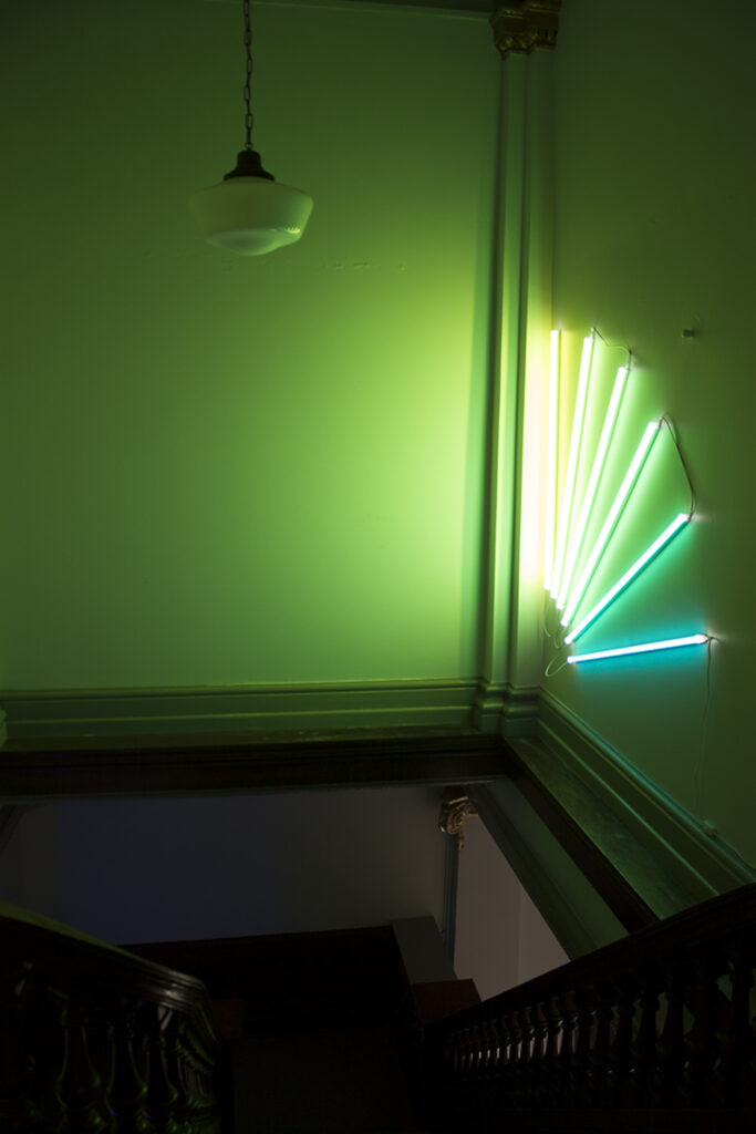 Image: An installation view of A Thought is a River, curated by John Knuth. Adrienne Dixon, Leur series, 2022, gels on LED, dimensions variable. A view looking down a staircase towards a corner, where a green neon installation is displayed. Photo by CM Turner.