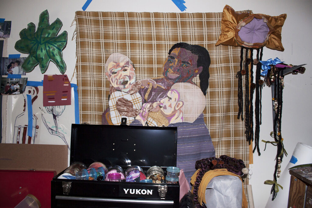 Image: An interior view of Josie Love Roebuck’s home/studio. Various textile works by the artist hang on the wall. The piece in the center depicts three figures interacting against a plaid background. Various art supplies are in the foreground. Photo by CM Turner.