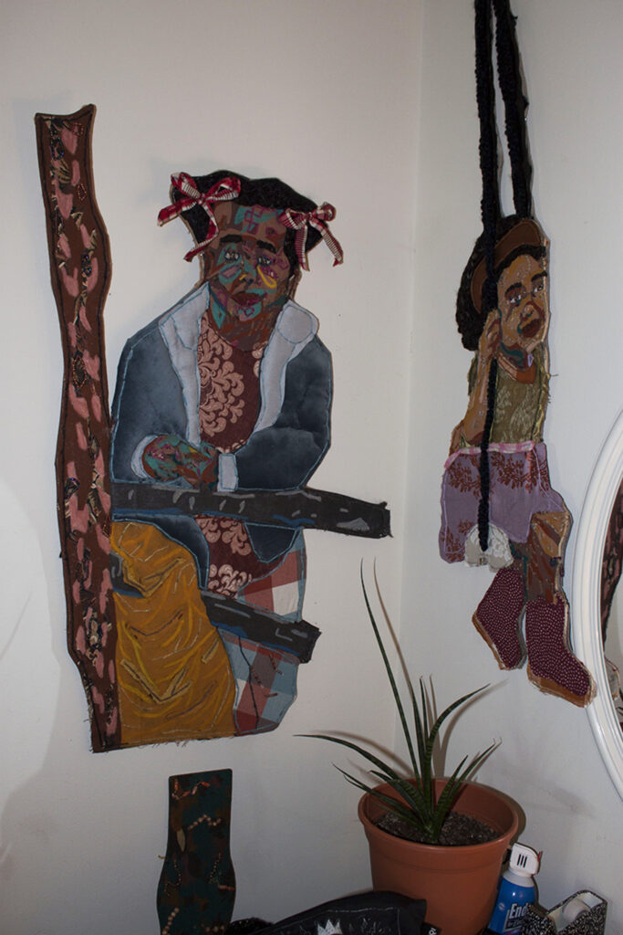 Image: An interior view of Josie Love Roebuck’s home/studio. Various textile works by the artist hang on the walls. The piece on the left depicts a Black person looking directly at the viewer. The piece on the right depicts a young Black girl on a swing. A plant is in the foreground. Image by CM Turner.