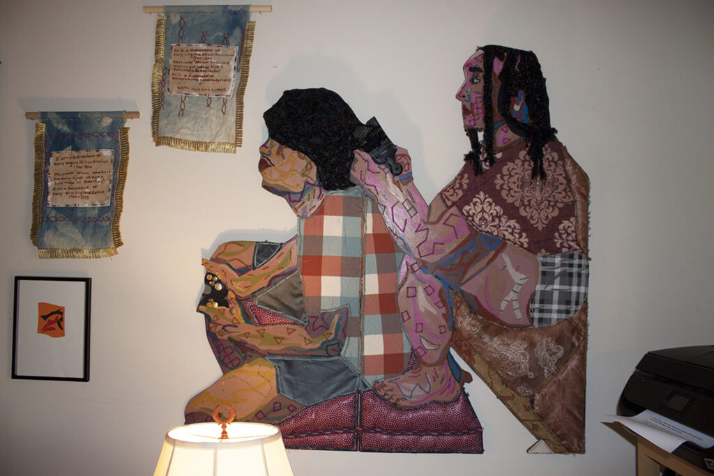 Image: An interior view of Josie Love Roebuck’s home/studio. Various textile works hang on the wall, with the primary piece depicting a figure sitting and doing the hair of another person siting next to them. Photo by CM Turner.