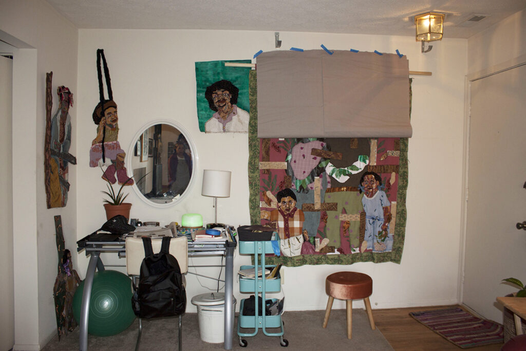 Image: An interior view of Josie Love Roebuck’s home/studio. Various textile works hang on the walls above and next to a table/work station with various art supplies. The large piece on the right depicts several people and patterns. Photo by CM Turner.