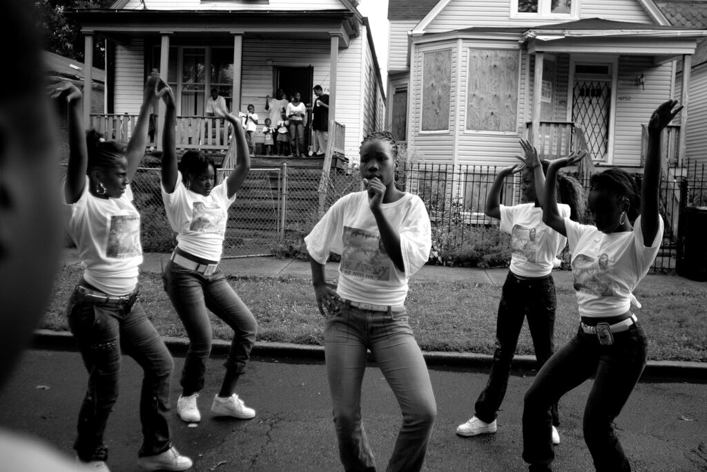 Image: A black and white photograph by Carlos Javier Ortiz. Englewood, Chicago, 2007. Ortiz: "Girls in the Englewood neighborhood on Chicago’s South Side attend a block party to celebrate the lives of Starkeisha Reed, 14, and Siretha White, 10. Starkeisha and Siretha were killed days apart in March 2006. The girls’ mothers were friends, and both grew up on Honore Street, where the celebration took place." The five girls are all wearing matching white shirts and dance in a formation. Image courtesy of the artist.