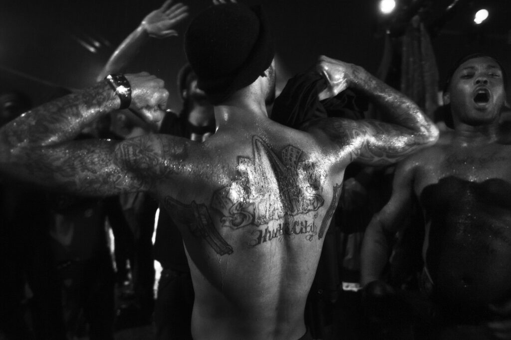 Image: Carlos Javier Ortiz, Mike Brown, 2014. Demonstrators block the streets in St. Louis to protest the killing of Michael Brown, 24 hours before a grand jury decided that police officer Darren Wilson would not face charges in the shooting of Michael Brown. Rioting and looting broke out soon after the announcement throughout the area leaving several businesses burned to the ground. The black and white photo shows a Black man facing away from the viewer. He has his arms up flexing. His arms and back have several tattoos. Image courtesy of the artist.