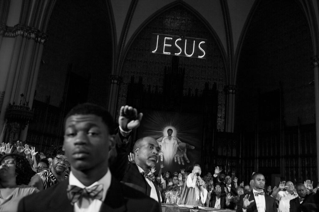Image: A black and white photo by Carlos Javier Ortiz. Auburn Gresham, Chicago, 2013. Ortiz: "Members of St. Sabina Church pray to end violence in Chicago. More than 50 young people have been murdered in the neighborhood since 2006." The photo shows a crowd in a church praying. There is a religious mural in the back with a neon sound reading "JESUS" above it. A man in the middle of the crowd has his hand in the air, as do many other people in the crowd. Image courtesy of the artist. 