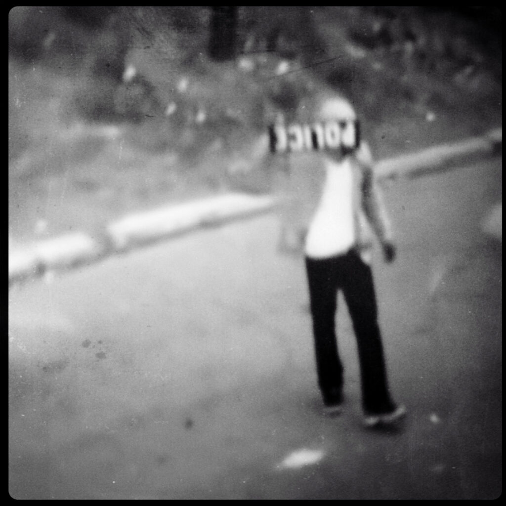 Image: A black and white photo by Carlos Javier Ortiz. Ortiz: "On April 12, 2015, Baltimore Police Department officers arrested Freddie Gray, a 25-year-old African American resident of Baltimore, Maryland. Gray sustained injuries to his neck and spine while in transport in a police vehicle. On April 18, 2015, after Gray's subsequent coma, the residents of Baltimore protested in front of the Western district police station. Gray died the following day, April 19, 2015, a week after the arrest. Further protests were organized after Gray's death became public knowledge, amid the police department's continuing inability to adequately or consistently explain the events following the arrest and the injuries. Spontaneous protests started after the funeral service, although several included violent elements. Civil unrest continued with at least twenty police officers injured, at least 250 people arrested, 285 to 350 businesses damaged, 150 vehicle fires, 60 structure fires, 27 drugstores looted, thousands of police and Maryland National Guard troops deployed, and with a state of emergency declared in the city limits of Baltimore. The state of emergency was lifted on May 6. On May 1, 2015, Gray's death was ruled by the medical examiner to be a homicide. Six officers were charged with various offenses, including second-degree murder, in connection with Gray's death. Three officers were subsequently acquitted; in July 2016, following the acquittals, Baltimore City State’s Attorney Marilyn Mosby dropped charges against the remaining three officers." A black and white, out of focus photograph of a person standing while holding a sign that reads "POLICE" upside down. Image courtesy of the artist.