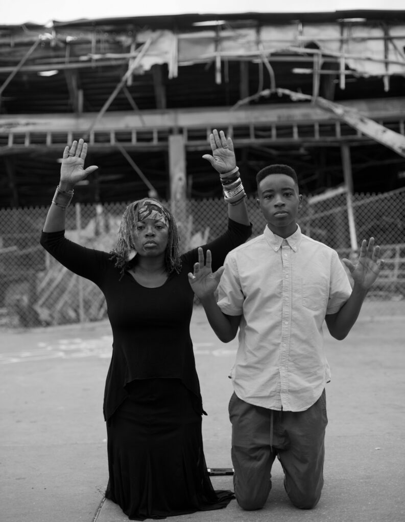 Image: Carlos Javier Ortiz, Poem to Benicio 2. A black and white photo of a Black woman and a Black man kneeling with their hands up. They are both directly facing the viewer. Image courtesy of the artist.